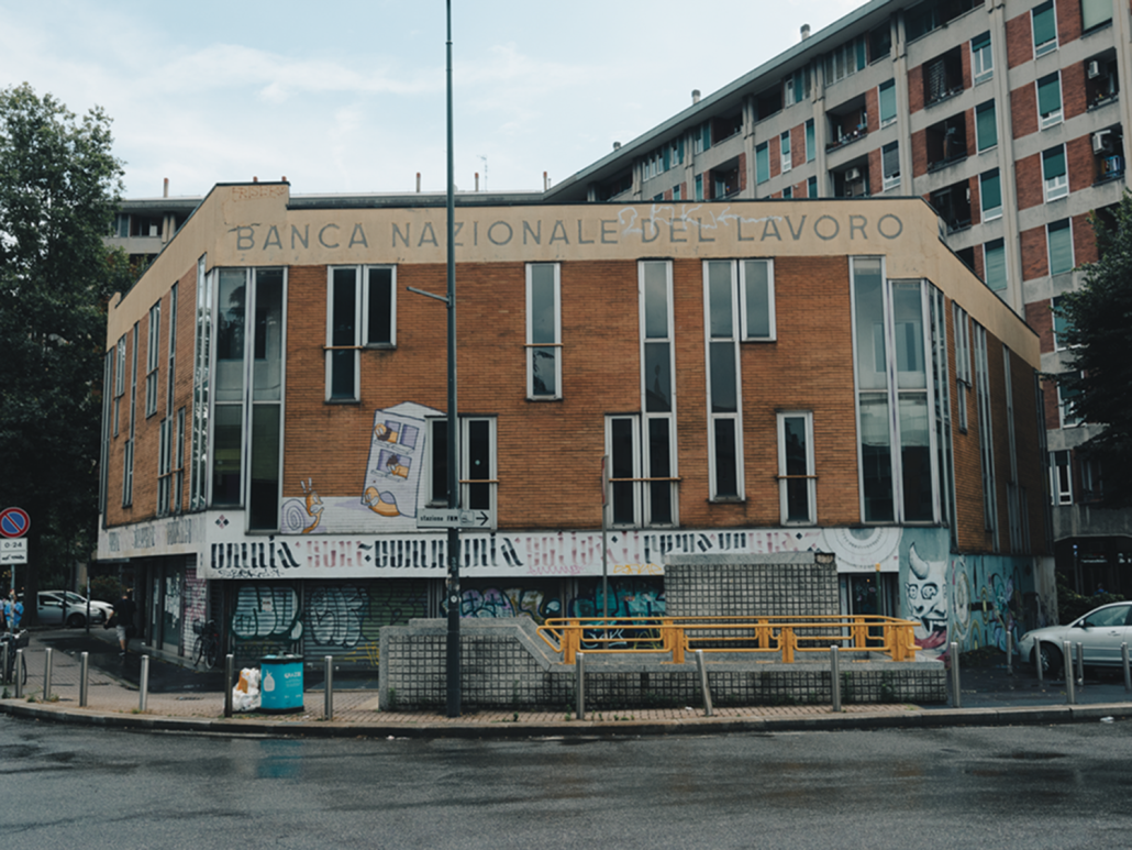 Alt: Mai+ Bologna - Opera dal titolo «1969 - liberamente ispirata» - foto dal vivo dell‘opera: si tratta di un‘incisione in intonaco bianco sul muro di un palazzo il cui intonaco di base è color terra bruciata, e ne compone una parte a mezzaluna sull’angolo del palazzo stesso